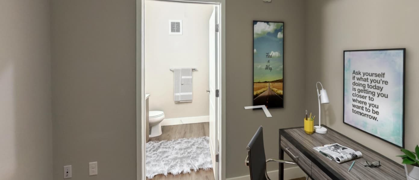 A modern home office with a desk, chair, lamp, and motivational wall art at Live at CoLab Denver; an open door reveals a bathroom with white walls, a towel rack, and a fluffy gray rug on the floor.