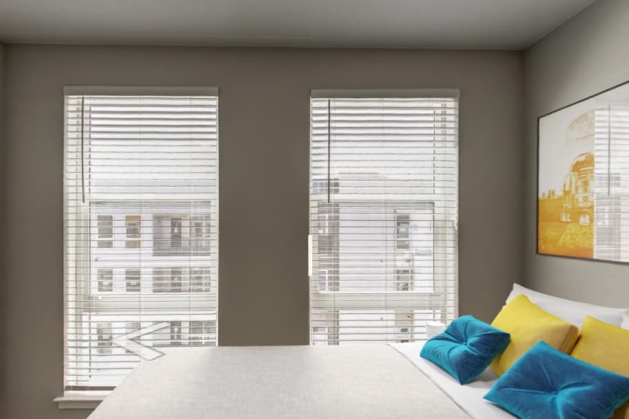 A modern bedroom at Live at CoLab Denver features two large windows with white blinds, a bed adorned with turquoise and yellow pillows, and a framed picture. White buildings are visible just outside the windows.
