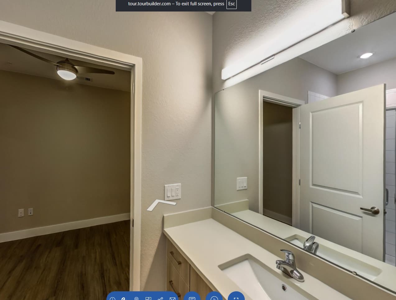 A modern bathroom with a large mirror, white countertop, and single sink. An open door leads to an adjacent room with wood flooring, beige walls, and a ceiling fan—experience stylish living when you Live at CoLab Denver.
