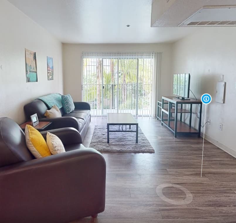 A bright living room at Aztec Corner Apartments features a brown leather sofa, armchair, coffee table, TV on a stand, and large windows with vertical blinds overlooking greenery. The wood floor and framed pictures add a cozy touch.