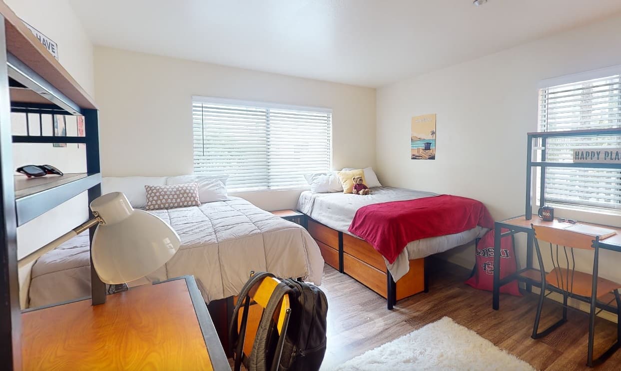 A tidy college dorm room at Aztec Corner Apartments with two beds, each with pillows and blankets, desks with lamps and chairs, a window with blinds letting in light, wooden floors, simple decor, and a backpack on a chair.