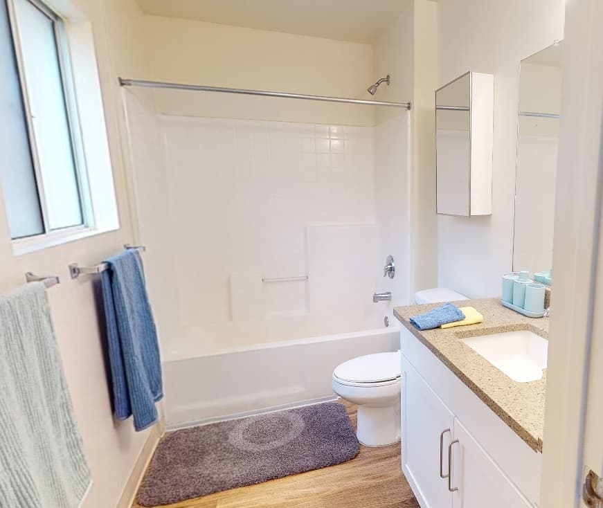 Modern bathroom at Aztec Corner Apartments with white walls, a bathtub with shower, toilet, white vanity and granite countertop. Towels hang by the window, and a gray bath mat sits on wood-style flooring—perfect student housing near campus.