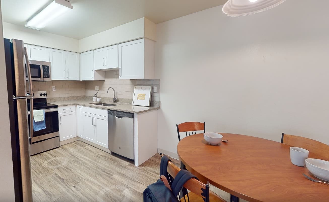 A modern kitchen at Aztec Corner Apartments features white cabinets, stainless steel appliances, wood-style flooring, and a round dining table for two. The bright, tidy space with neutral tones is perfect for student housing near campus.