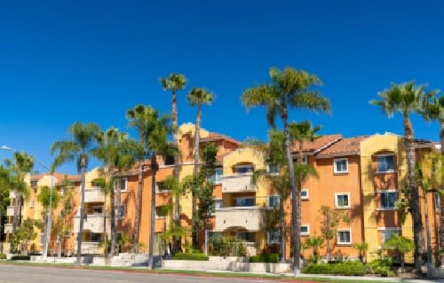 A modern orange apartment building, Aztec Corner Apartments, features balconies lined with tall palm trees under a clear blue sky.