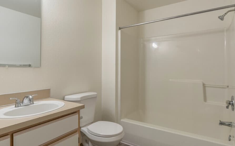 A clean, simple bathroom at Crystal Lake Apartments features a white sink, countertop, toilet, and a bathtub-shower combo with a shower curtain rod. Light-colored walls and a wall-mounted mirror complete the space.