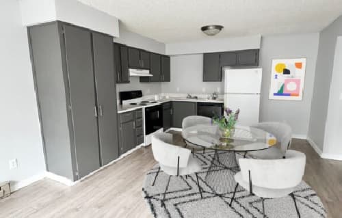 Modern kitchen and dining area at Cougar Ridge Apartments features gray cabinets, white appliances, a round glass table with four light gray chairs, patterned rug, and abstract wall art. Light wood flooring and neutral walls complete the space.