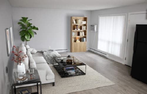 A modern living room at Cougar Ridge Apartments features a white sectional sofa, glass coffee table, bookshelf with books and decor, black refrigerator, large windows with sheer curtains, and a tall green plant in the corner.