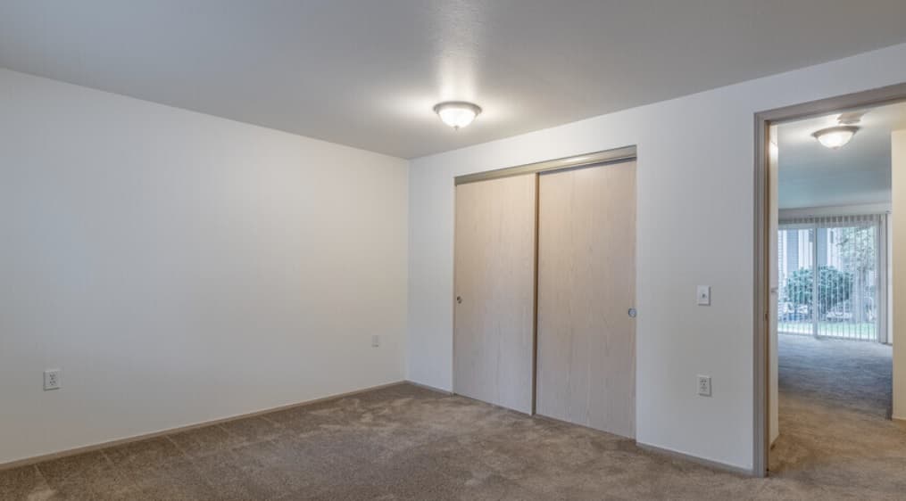 Empty bedroom at Crystal Lake Apartments with beige carpet, white walls, a ceiling light, and a closed double-door closet. An open doorway leads to a brightly lit room with vertical blinds and outdoor views.