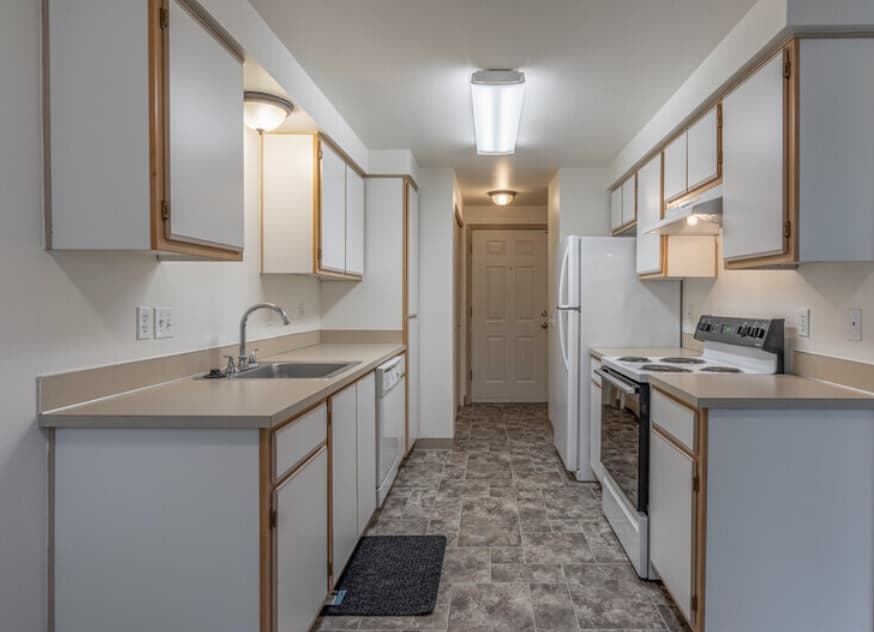 A narrow kitchen at Crystal Lake Apartments with white cabinets and light countertops features a sink, dishwasher, stove, oven, and refrigerator. The tiled floor and overhead lights brighten the space, with a door visible at the end of the hallway.