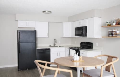 Modern kitchen at Cougar Ridge Apartments with white cabinets, black appliances, sink, and open shelves. A round wooden dining table for two is set with decorative items, complementing the neutral and minimalist color scheme.