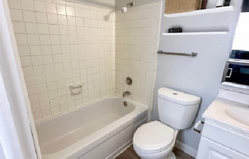 A small bathroom at Cougar Ridge Apartments features a white bathtub and tiled shower, a white toilet, shelves with storage, and a white sink vanity set against light gray walls.