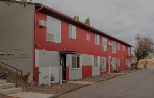 Cougar Ridge Apartments is a two-story red and gray building featuring several windows and air conditioning units, set beside a paved roadway with scattered trees and cloudy skies overhead.
