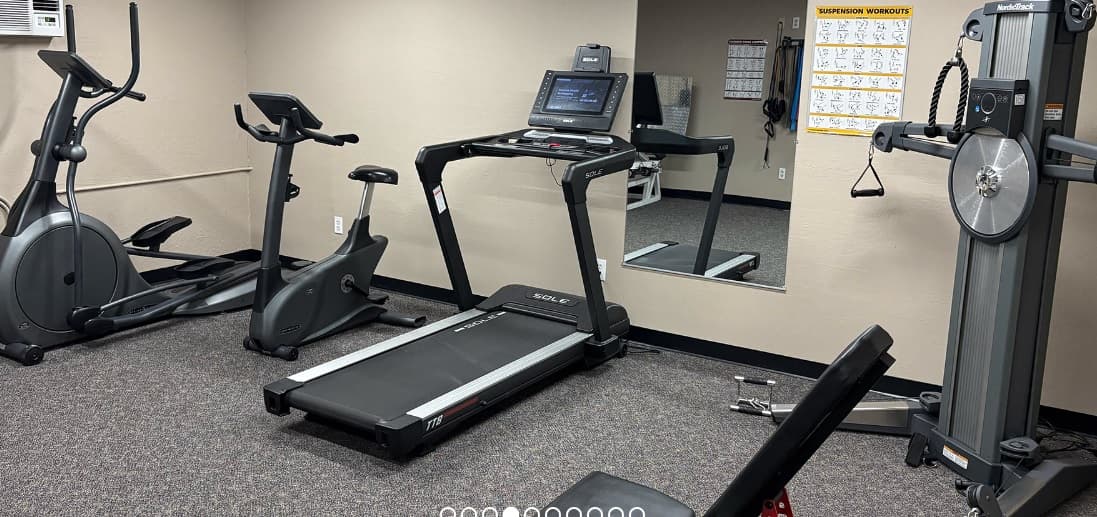A small gym room at College Terrace Apartments Reno features a treadmill, stationary bike, elliptical, weight machine, bench, and mirrored wall. An exercise chart is mounted on a tan wall above the carpeted flooring.