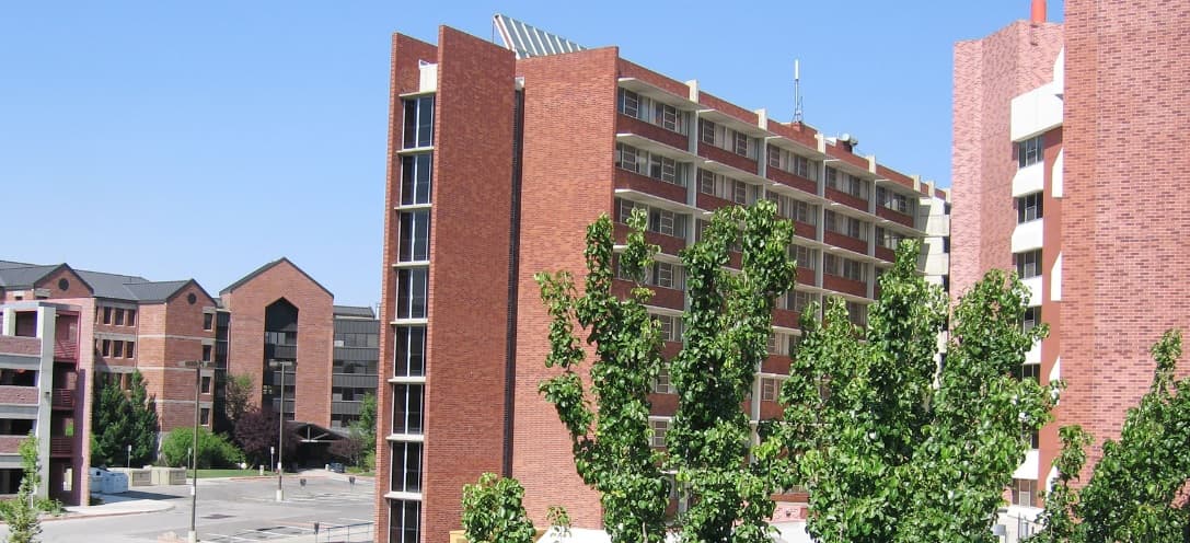 The College Terrace Apartments Reno features multi-story red brick buildings with balconies, set against a clear blue sky, with green leafy trees in the foreground and a parking lot visible to the left.