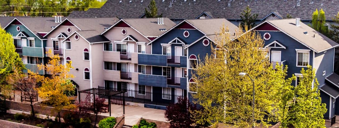 A row of modern, multi-story College Terrace Apartments Reno buildings with balconies, colorful siding, and decorative gables, surrounded by lush green trees and landscaping in spring.
