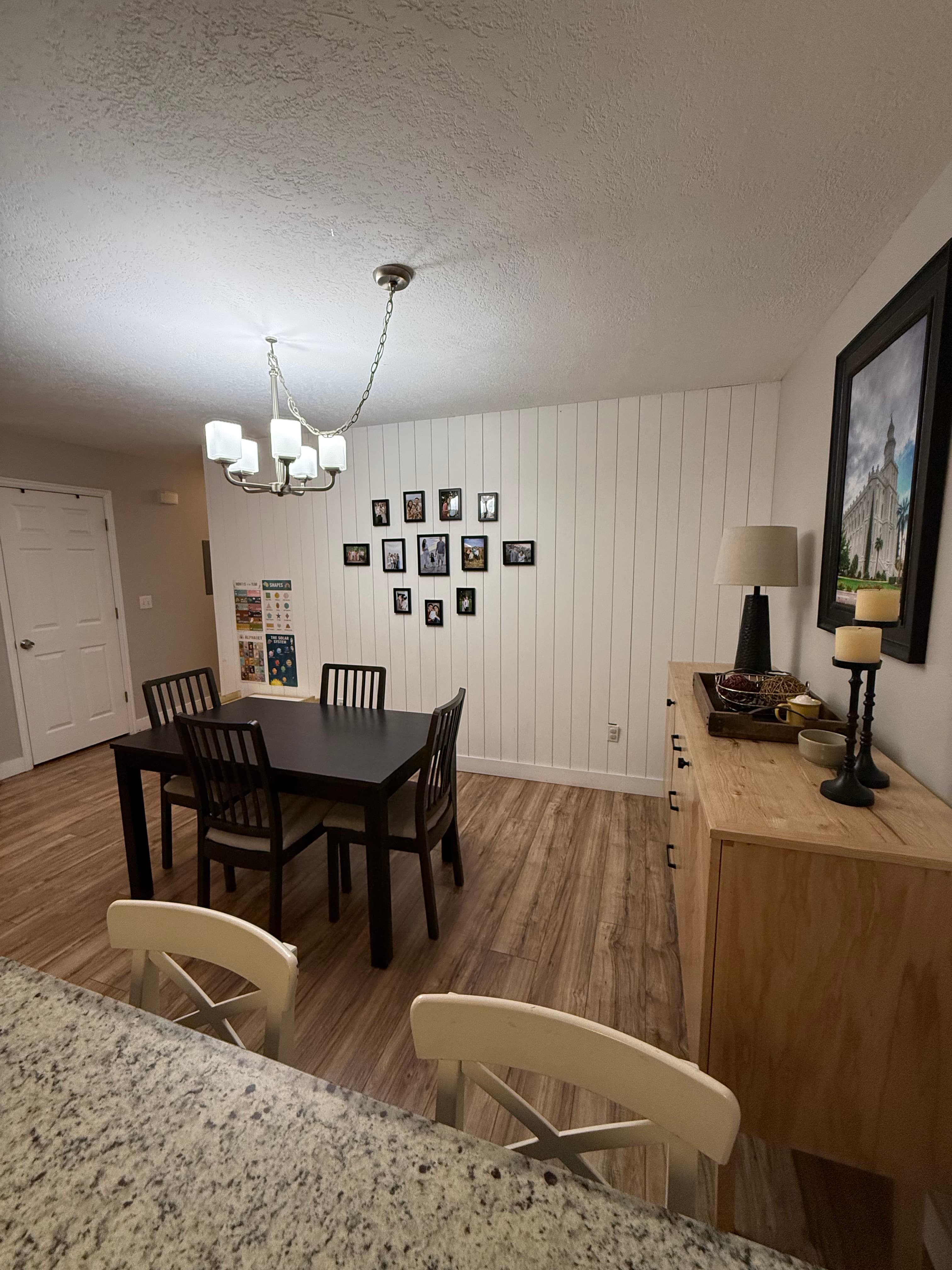 A dining area at 282 W 960 S, APT C features a dark table with six chairs on wood flooring. A chandelier hangs overhead, a sideboard with decor lines the right wall, and a gallery of framed photos adorns the back wall.
