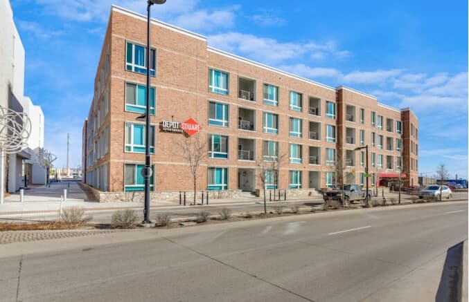 A modern, three-story brick apartment building with balconies and large windows stands along a clean, empty street under a bright blue sky. A sign on the corner reads Depot Square Apartments Boulder.
