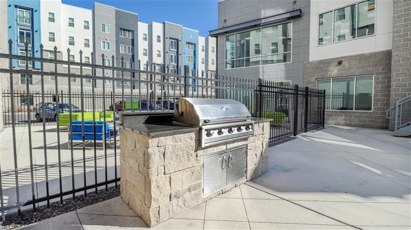 An outdoor stainless steel gas grill built into a stone counter sits on a concrete patio near a black metal fence at Promenade Place Orem, with modern apartment buildings and patio furniture in the background.