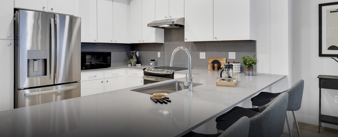 Modern kitchen with white cabinets, stainless steel appliances, a gray backsplash, and a large island with a sink, faucet, and three barstools. Countertops feature minimal decor and small kitchen essentials.