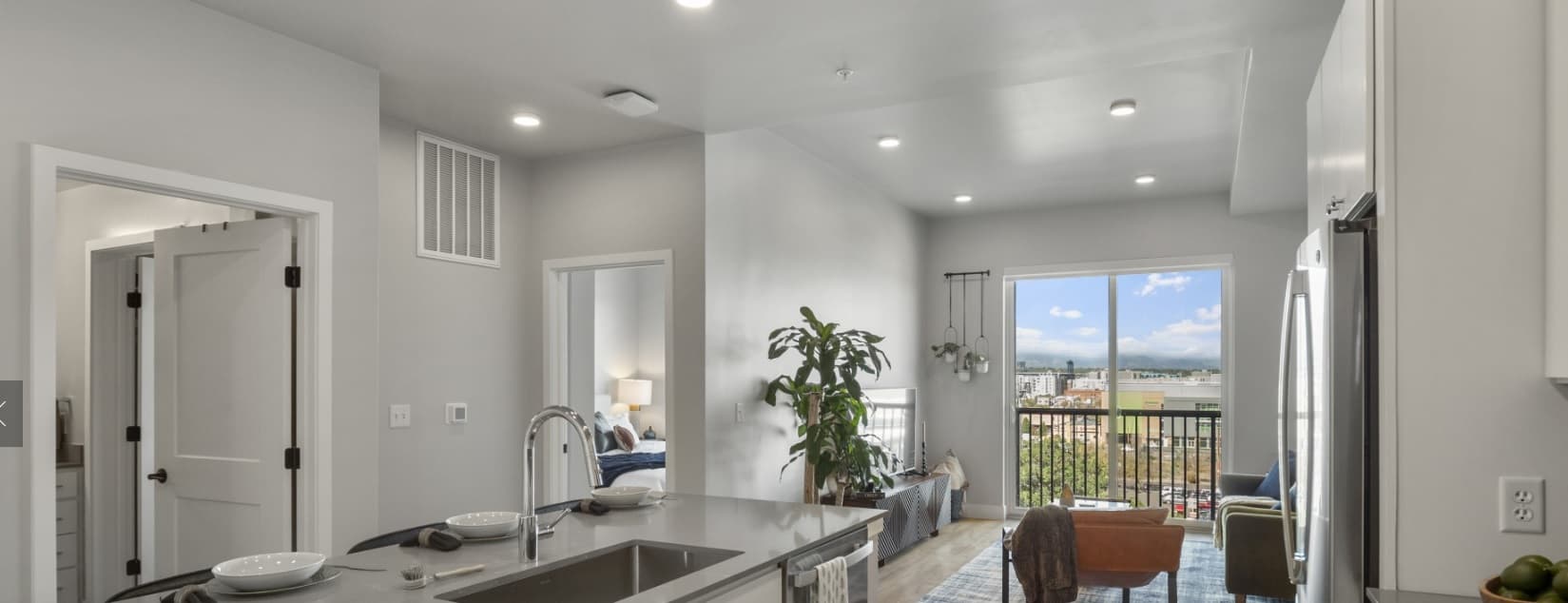 Modern apartment interior with a kitchen island in the foreground, a living area with a couch and plant, large windows letting in natural light, and an open bedroom door visible on the left.