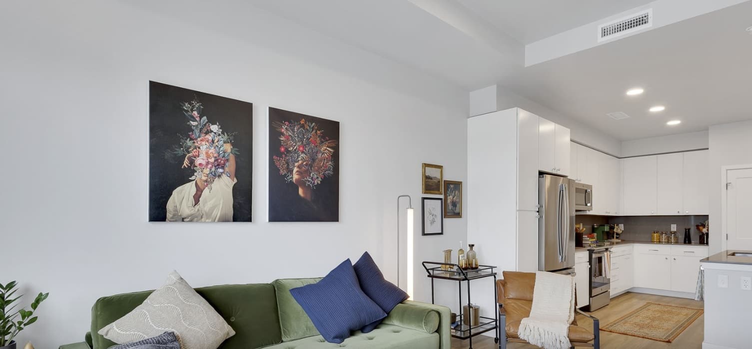 A modern living room with a green sofa, blue and beige pillows, wall art featuring floral portraits, and a view into a white kitchen with stainless steel appliances. A tall lamp and wooden chair are also visible.