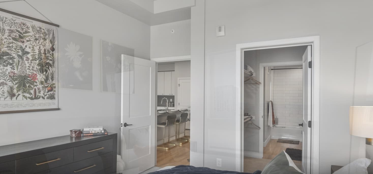 A modern bedroom with light gray walls, dark dresser, botanical wall art, and a partially open door leading to a kitchen. Another door reveals a walk-in closet and a bathroom with a glass shower.
