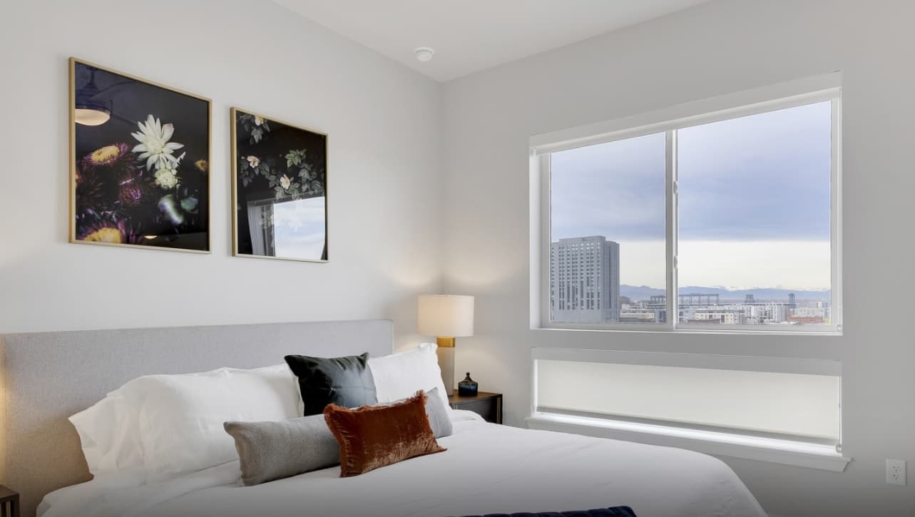 A modern bedroom with a large bed, white bedding, and decorative pillows. Two floral art pieces hang above the bed. A window shows a cityscape with tall buildings and mountains in the distance.