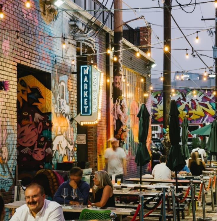Outdoor dining area with string lights, murals on brick walls, people seated at tables, and a neon Market sign at dusk, creating a lively urban atmosphere.