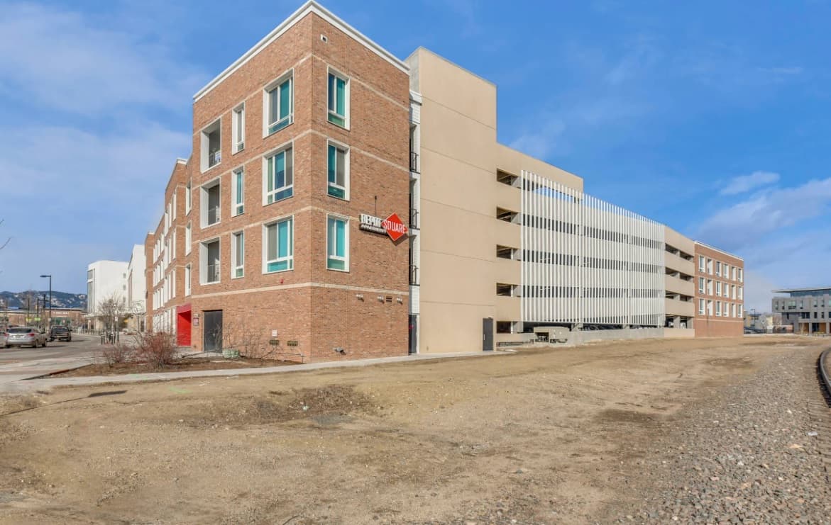 A modern red-brick building with red-trimmed windows, part of Depot Square Apartments Boulder, sits next to a multi-level parking garage and railroad tracks under a clear blue sky. The ground in front is bare dirt and gravel.