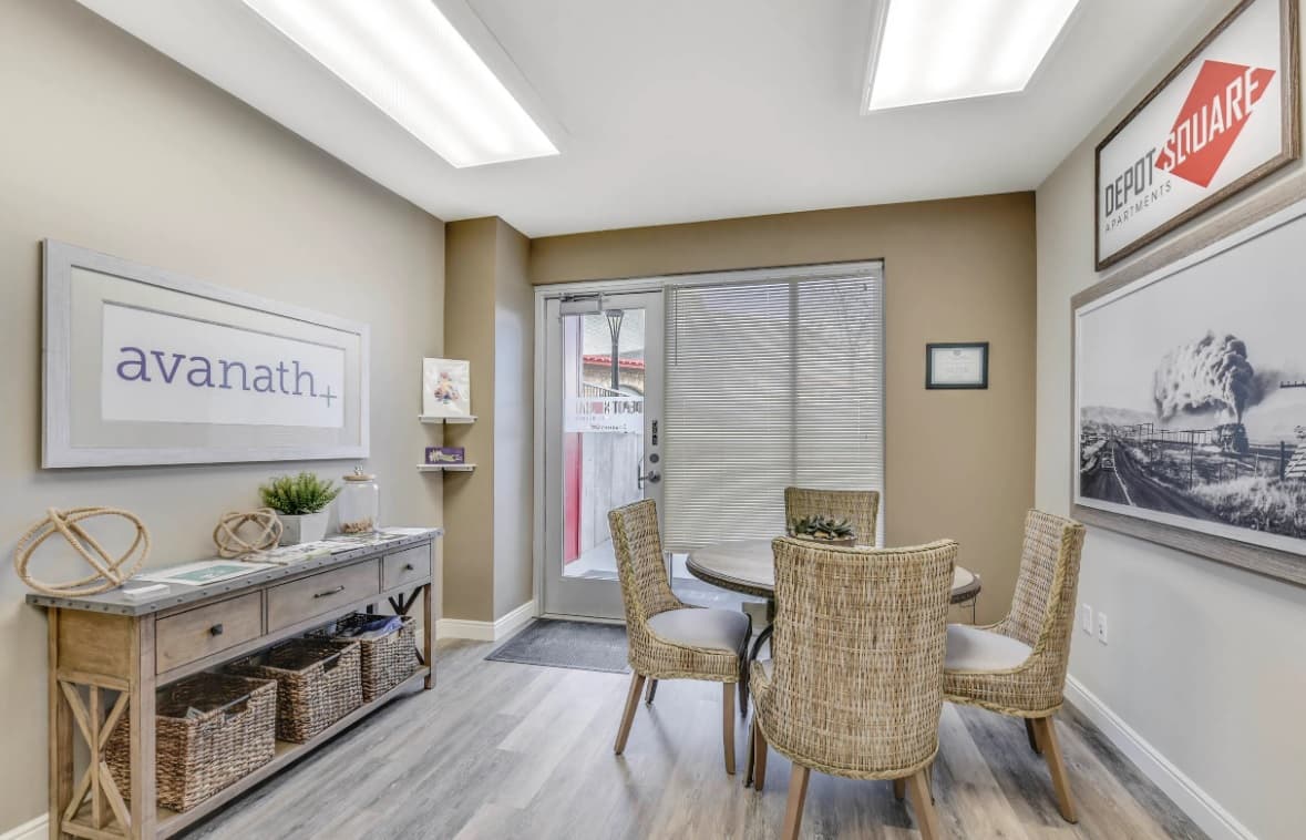 A small office at Depot Square Apartments Boulder features a round table with four wicker chairs, a console table with baskets and decor, wall art, and a glass door with blinds opening outside. Neutral colors and bright ceiling lights complete the space.
