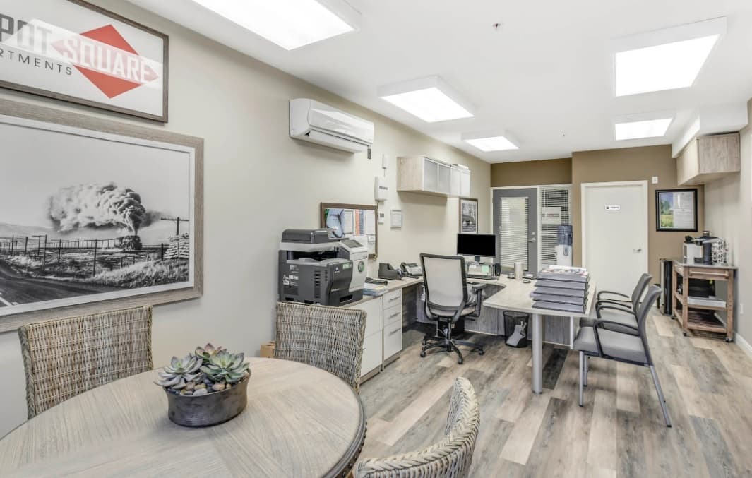 Modern office at Depot Square Apartments Boulder with desks, chairs, computers, and a printer. A round table with wicker chairs and a small succulent plant sits in the foreground. Neutral colors and framed art create a clean, organized workspace.