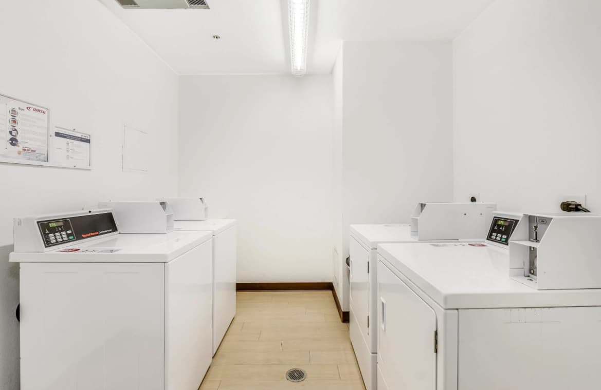 A bright, clean laundry room at Depot Square Apartments Boulder with four white, front-loading washing machines and dryers arranged in a corner, light wood flooring, and white walls with instructional signs.