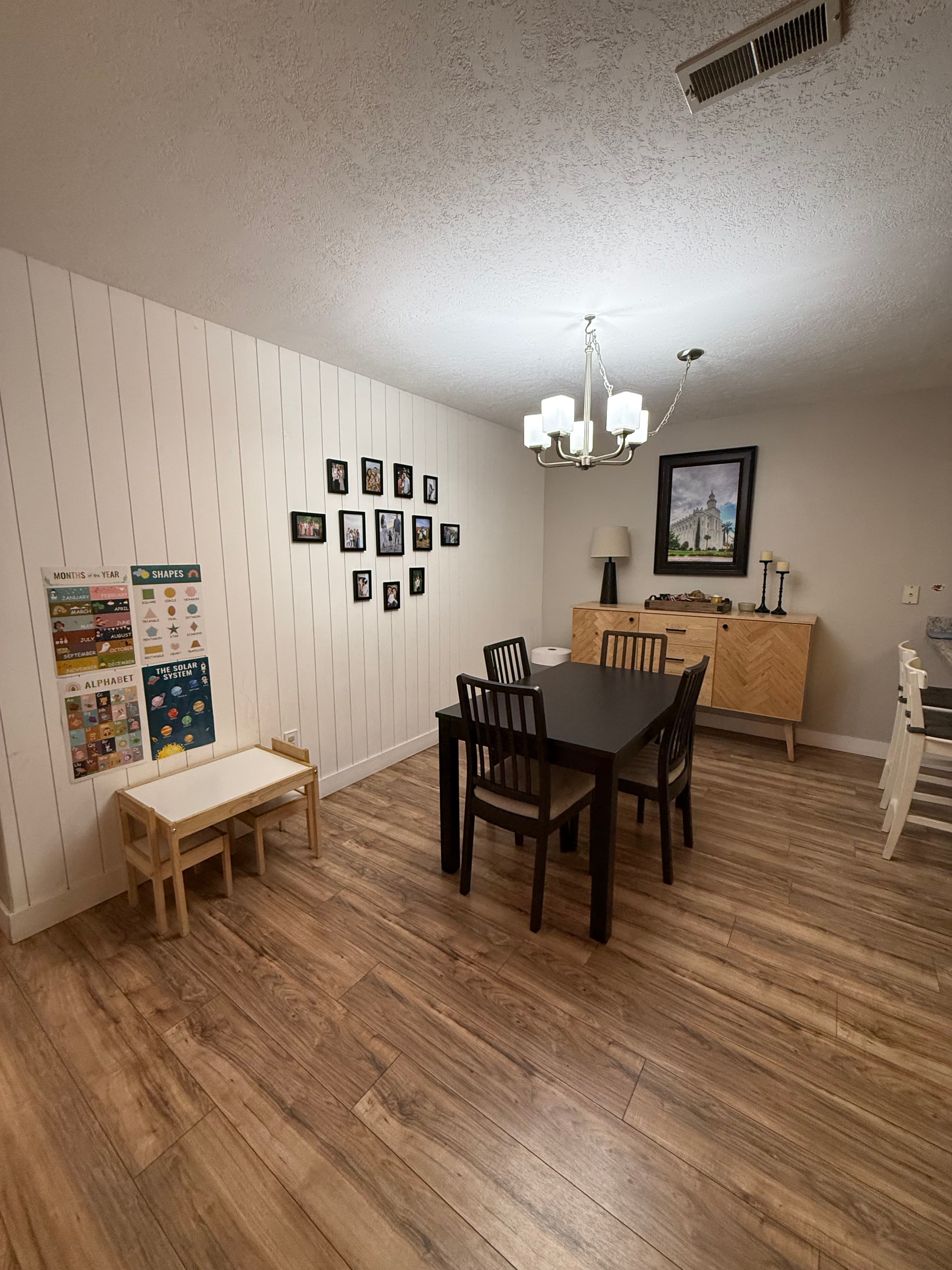 A stylish apartment dining room at 282 W 960 S features a black table with four chairs, wooden floors, white paneled walls, a chandelier, wall art, framed photos, a sideboard, children’s bench, and educational posters on the left wall.