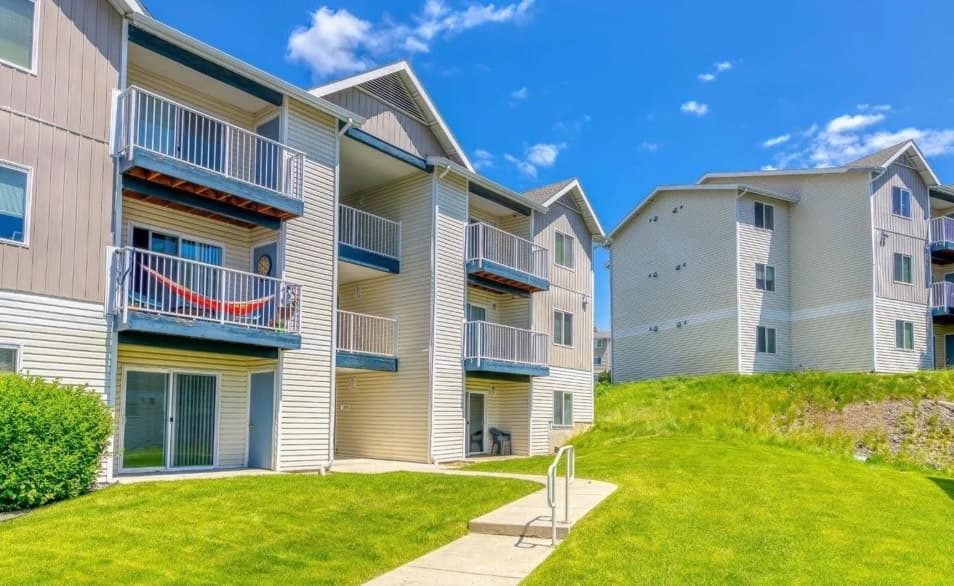 Two light-colored apartment buildings with balconies sit under a bright blue sky at Emerald Downs Apartments. A sidewalk leads to one entrance, and green grass surrounds the buildings with some bushes and a grassy slope.