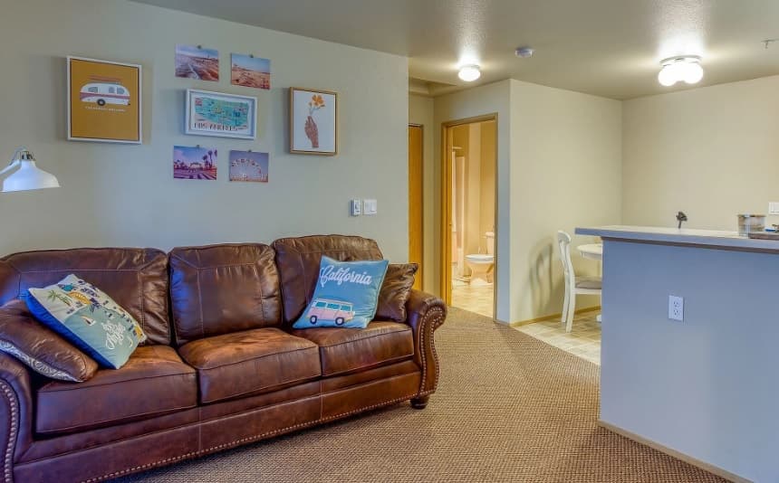 A cozy living room at Emerald Downs Apartments features a brown leather sofa, colorful travel-themed pillows, and framed art. The space opens to a small dining area and kitchen with warm lighting and neutral walls and carpet.