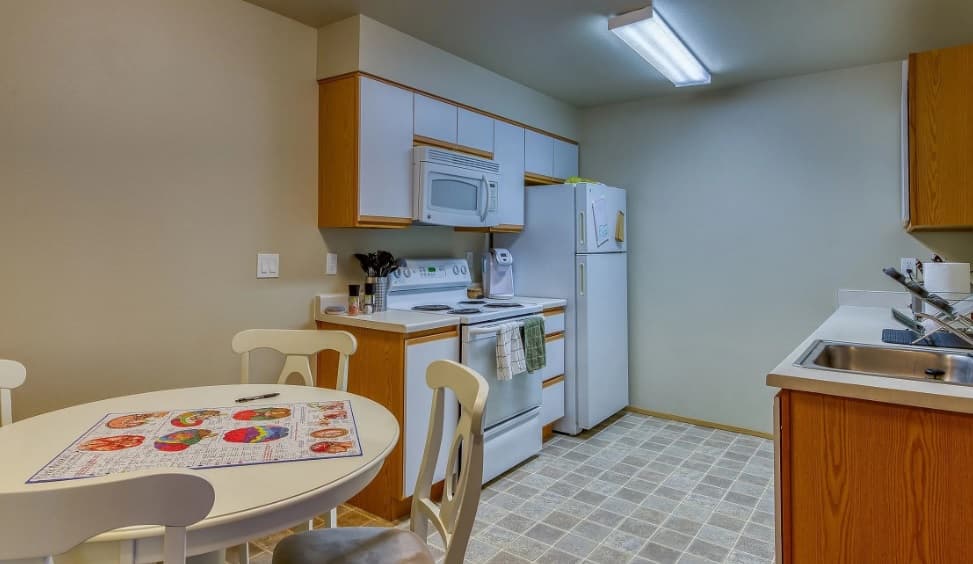 A small kitchen at Emerald Downs Apartments features light wood cabinets, white appliances, and a round dining table with a colorful placemat. Tiled floors and a welcoming layout make this space ideal for comfortable apartment living.