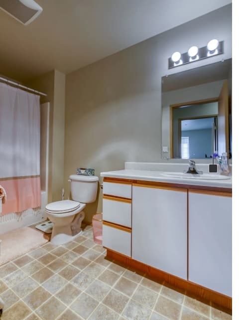 A clean bathroom at Emerald Downs Apartments features a white vanity, large mirror, sink, and overhead lights. There’s a toilet, a shower with a curtain, beige tile flooring, plus toiletries and decor on the counter and floor.