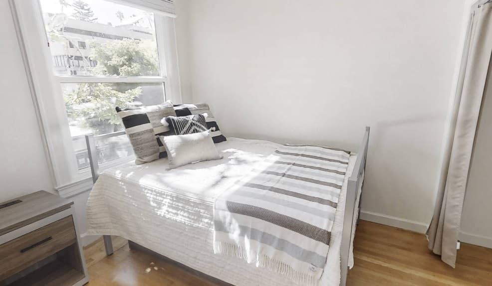A neatly made bed with striped pillows and a blanket is placed beside a large window letting in natural light. The room has light-colored walls, wooden flooring, and a small brown nightstand next to the bed.