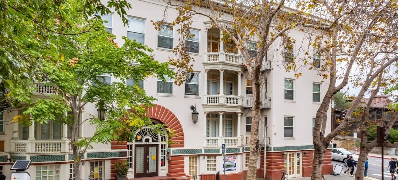 A four-story apartment building with white walls, red brick accents, arched entryway, balconies, and large windows. Leafy trees partially obscure the facade, and people stand near the entrance on the sidewalk.