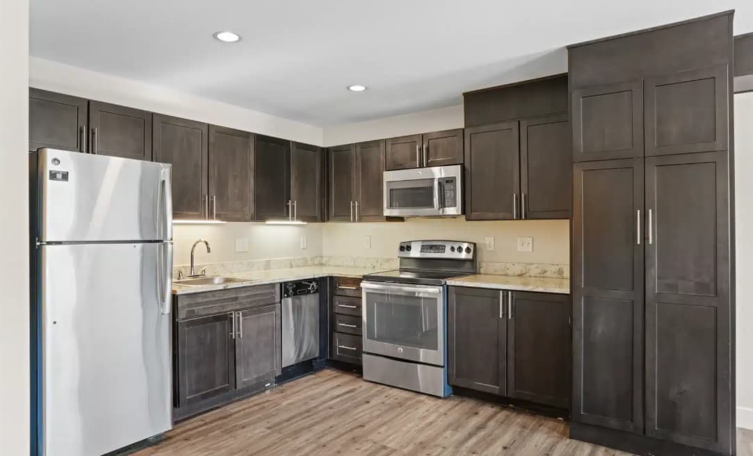 Modern kitchen at East Village Flats Boulder with dark wood cabinets, stainless steel appliances including a refrigerator, stove, oven, microwave, and dishwasher, light countertops, wood-style flooring, bright lighting, and neutral wall colors.