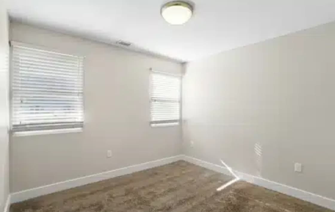 Empty room with beige carpet, light walls, and two windows with white blinds at East Village Flats Boulder. Sunlight streams through the blinds, creating light patterns on the carpet beneath the ceiling light fixture.