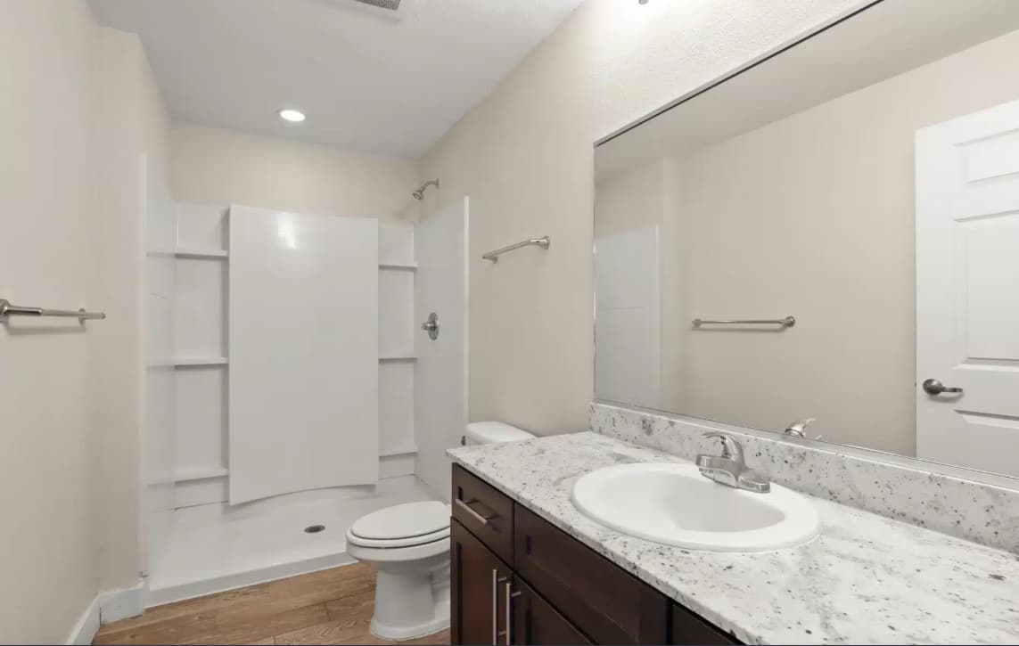 A modern bathroom at East Village Flats Boulder features beige walls, a large mirror above a granite countertop with sink, dark wood vanity, toilet, and a white walk-in shower with built-in shelves and a silver showerhead.