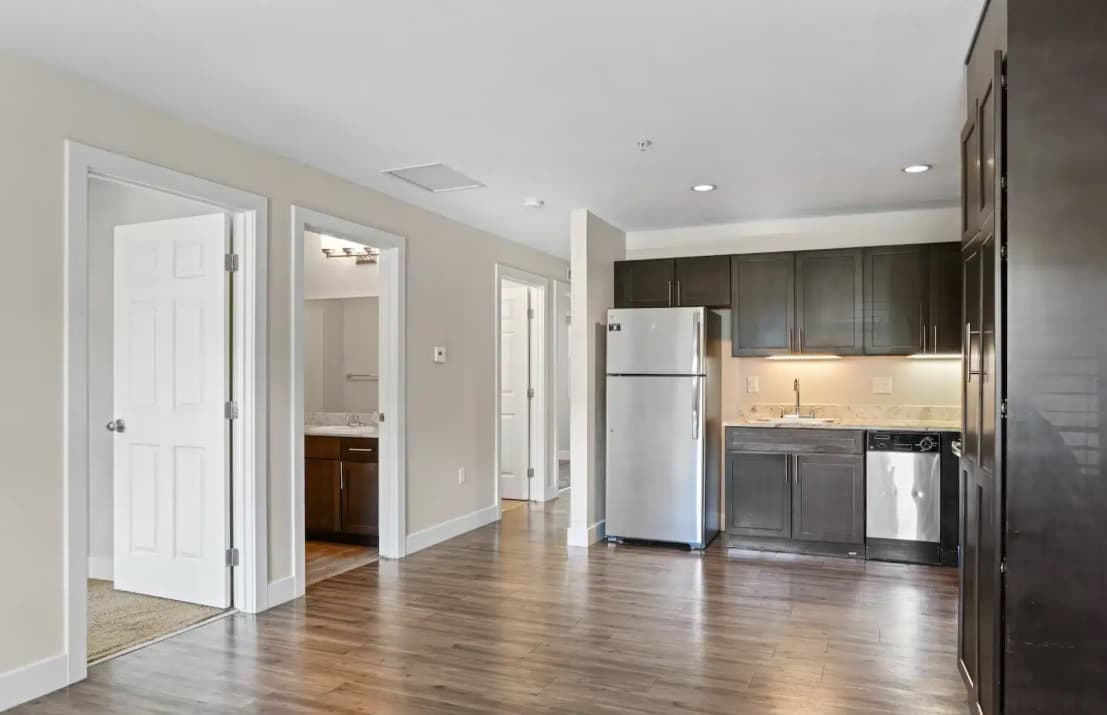 Modern East Village Flats Boulder apartment kitchen with stainless steel appliances, dark cabinets, wood flooring, and beige walls. Open layout connects to a hallway with white doors leading to a bathroom and another room.