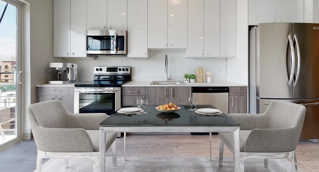 Modern kitchen at Evo on Emerson with white and gray cabinets, stainless steel appliances, a dining table set for two with gray chairs, and a bowl of fruit in the center. Large window provides natural light.