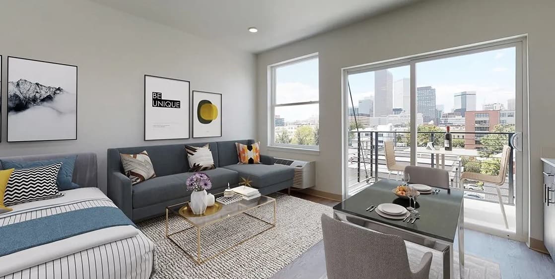 Modern studio apartment at Evo on Emerson with a bed, grey sofa, coffee table, and dining table set for two. Large windows and a door open to a balcony with city skyline views. Walls feature minimalist artwork.