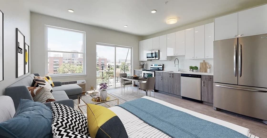Modern studio apartment at Evo on Emerson with a bed, sofa, and small dining table in an open space. Large windows let in natural light, and a compact kitchen features stainless steel appliances and white cabinets. City buildings visible outside.
