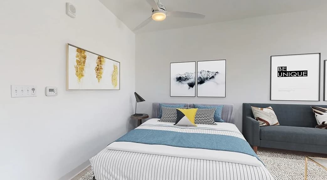 A modern bedroom at Evo on Emerson with a bed featuring striped bedding and colorful pillows, a gray sofa, abstract wall art, a floor lamp, and a ceiling fan. The minimalist decor has white walls and neutral tones.