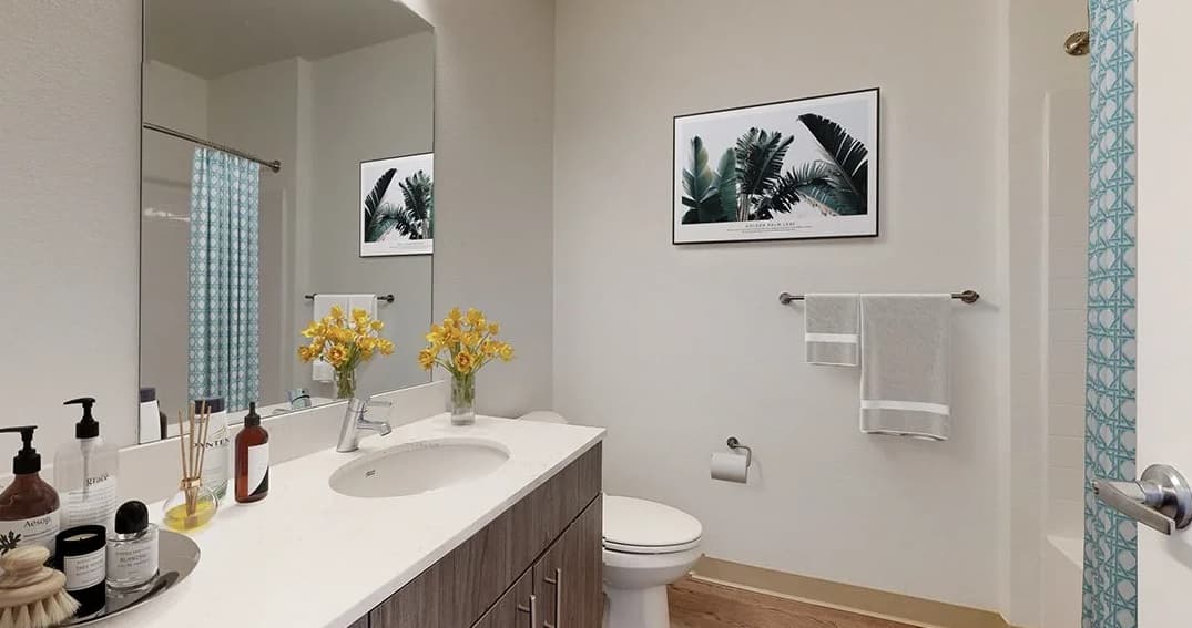 Modern bathroom at Evo on Emerson with a large mirror, white countertop, wooden vanity, yellow flowers in vases, toilet, palm leaf wall art, and teal-patterned shower curtain. Towels hang on the wall and counter.