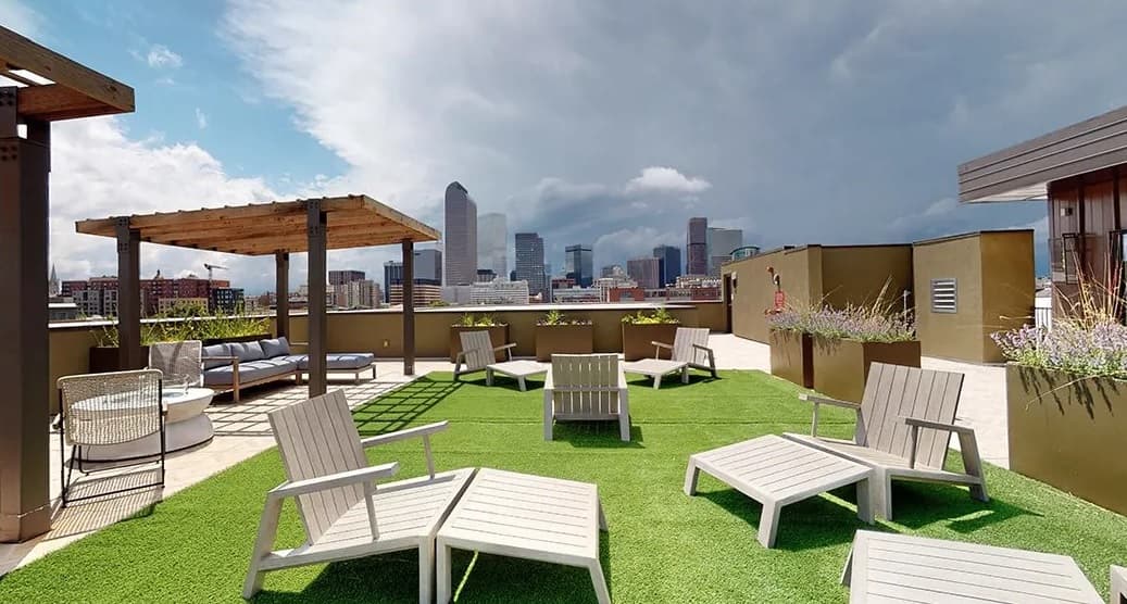 Rooftop lounge at Evo on Emerson with artificial grass, white lounge chairs, a wooden pergola over a cozy seating area, potted plants, and stunning city skyline views beneath a partly cloudy sky.