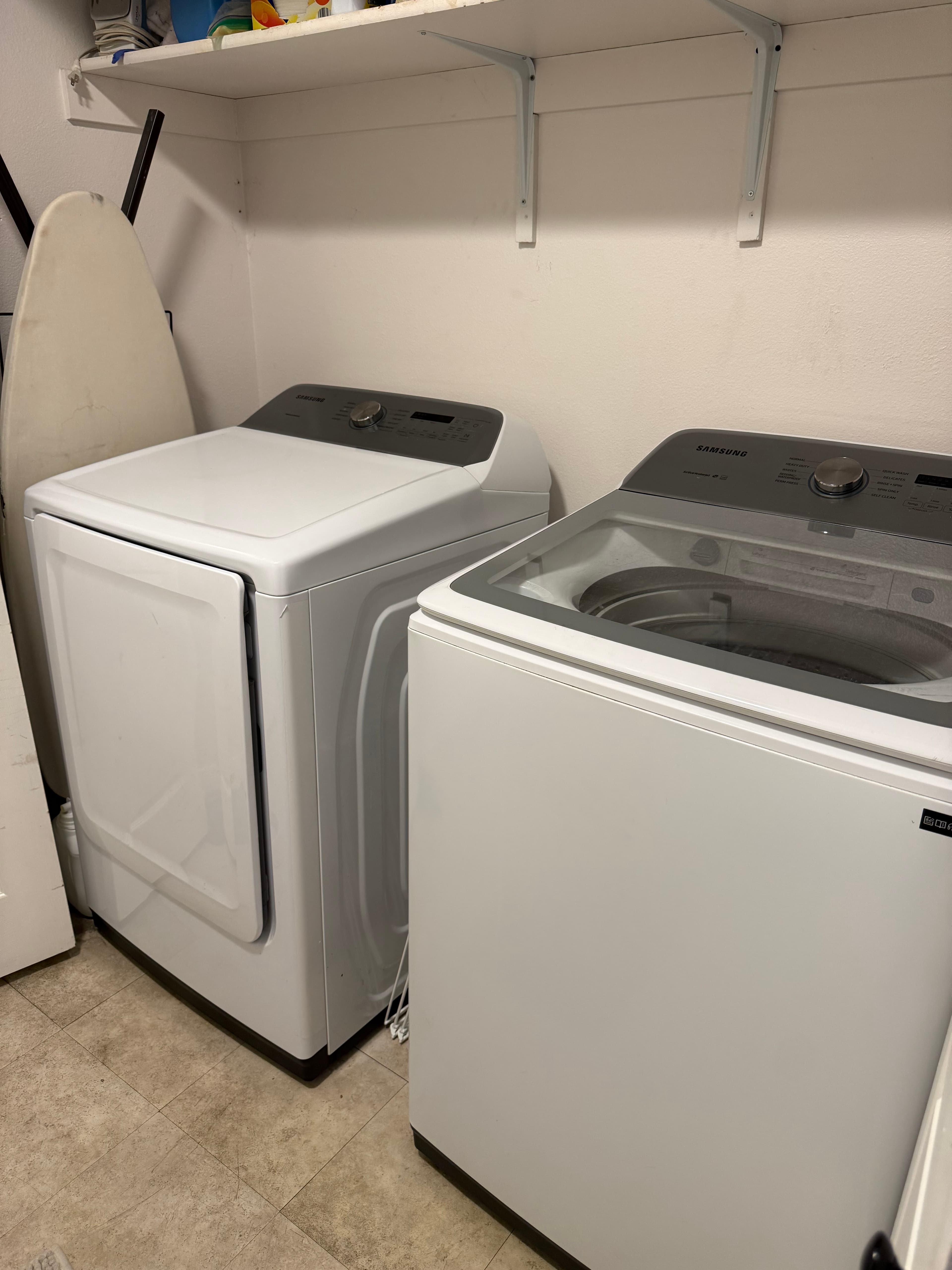 A laundry room in APT C, 282 W 960 S features a white front-loading dryer, a top-loading washing machine, an ironing board, and white shelves holding various items against a clean white wall.